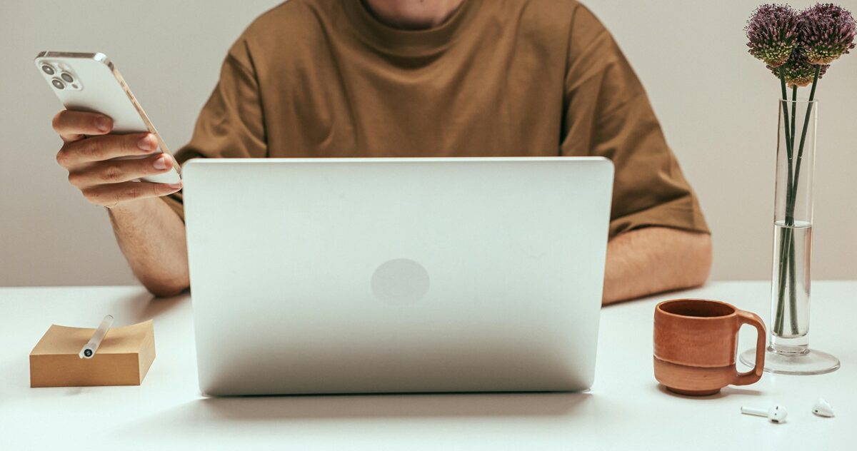 Person working from home on a laptop while holding a smartphone, with a mug and flowers on the desk